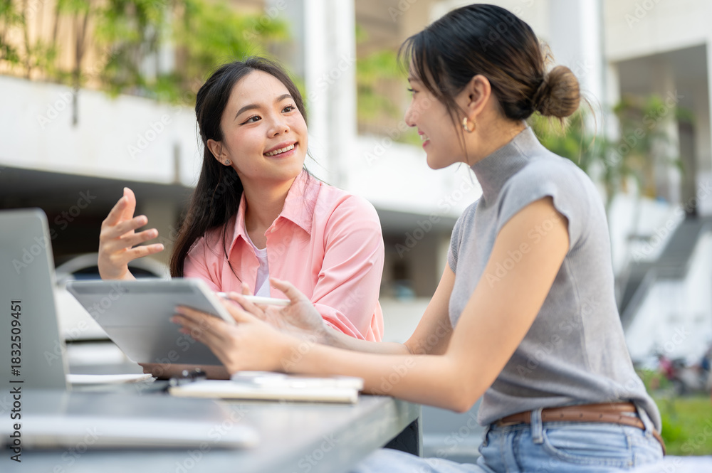 Fototapeta premium Asian woman student talking to friend holding tablet working on group project at table in university