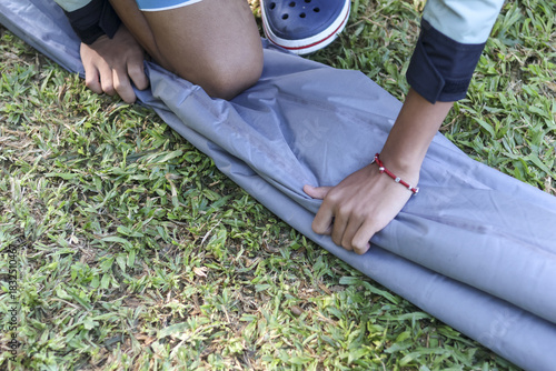 Focused person folding and packing an air mattress outdoors on green grass. Camper hand rolling grey sleeping pad after camping trip, preparing for journey home