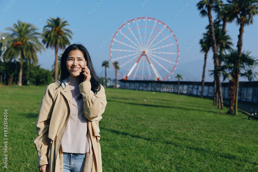 Fototapeta premium Woman in a beige coat smiles while talking on her phone in a sunny park, green grass and a distant ferris wheel creating a casual outdoor lifestyle scene