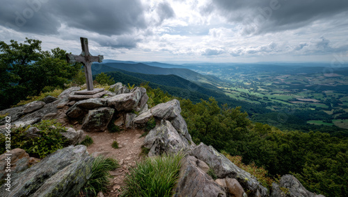 Cross atop a rocky peak overlooking a vast green valley under a cloudy sky.