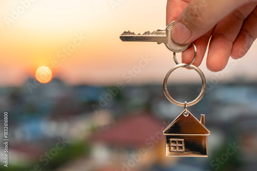 Hand holding a house keychain with a key against sunset sky, symbolizing new home ownership, real estate success, and fresh beginnings. Perfect for New Year goals, property, and investment concepts.