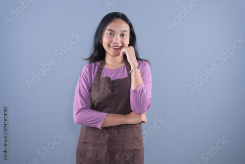 A smiling woman in a brown apron is posing with her hand on her chin
