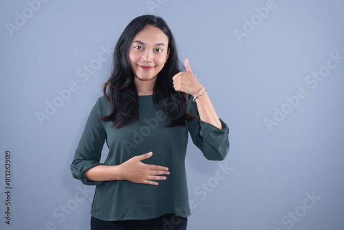 A smiling woman in a dark green top gives a thumbs up and holds her stomach