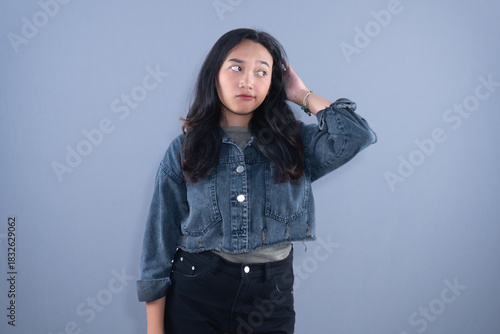 A young woman in a denim jacket looks away with a thoughtful or confused expression, hand on head