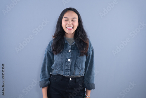 A young woman in a denim jacket smiles with eyes closed, sticking her tongue out playfully