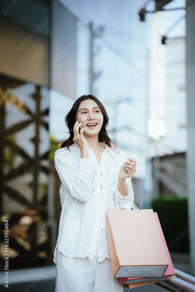 Fototapeta premium An Asian woman happily talking on the phone while holding shopping bags outside a modern building, capturing the joyful lifestyle of a young Asian woman on a shopping day.