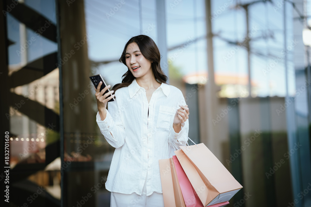 Fototapeta premium An Asian woman happily talking on the phone while holding shopping bags outside a modern building, capturing the joyful lifestyle of a young Asian woman on a shopping day.