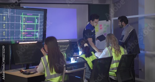 Engineers in high visibility vests collaborate in control room, reviewing digital site plans and dashboards during an operations and safety discussion. Coordination, monitoring, and incident readiness