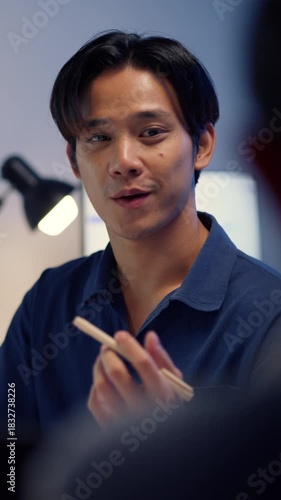 A close-up portrait of a serious Asian businessman discussing or presenting project details to a colleague while working late in the office. The desk lamp provides focused illumination.