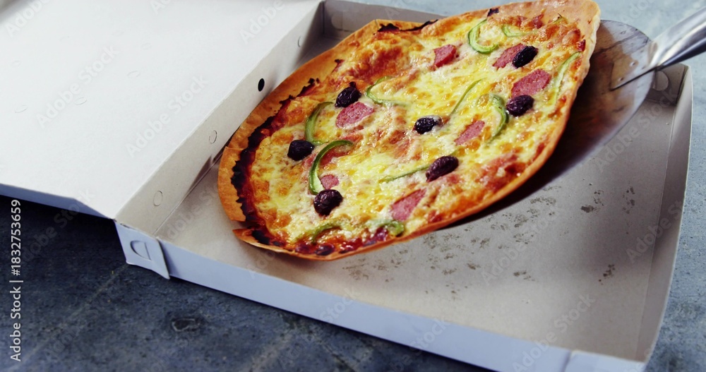Fototapeta premium Lifting thin-crust pizza on metal spatula in white box on gray tabletop, showing pepperoni, olives