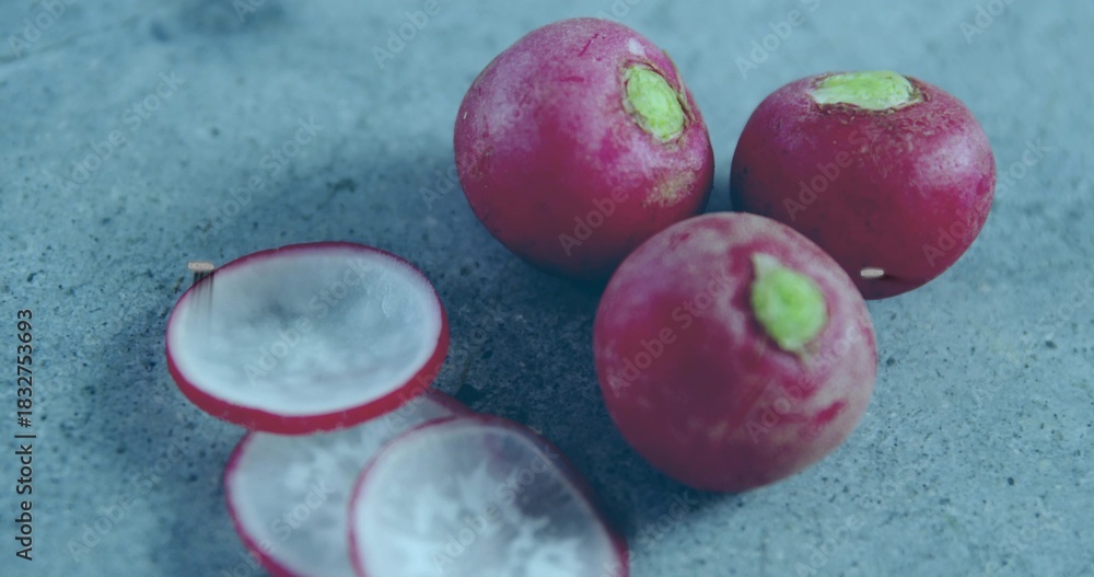 Fototapeta premium Displaying three whole radishes with trimmed green tops and two slices on bluish-gray stone surface