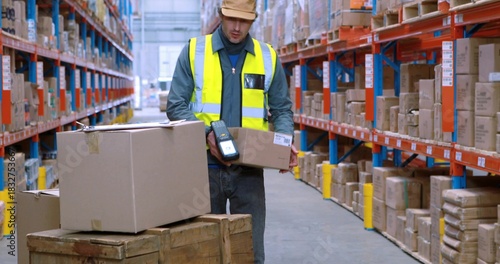 Scanning worker wearing hi-vis vest and cap, operating scanner in warehouse, with pallets