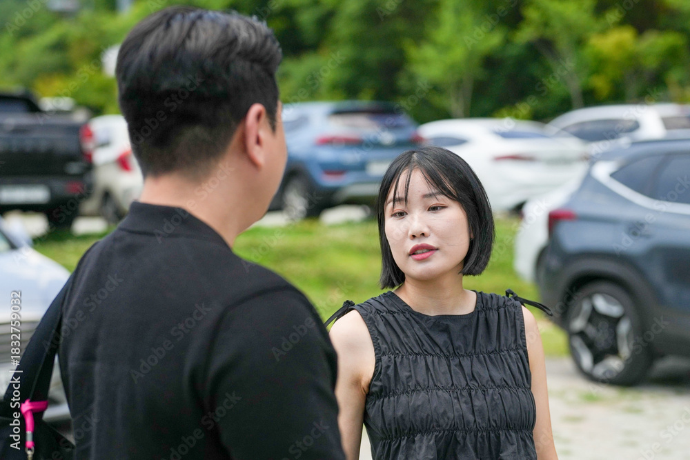 Obraz premium A man and a woman are talking to each other in a parking lot. The woman is wearing a black dress and the man is wearing a black shirt. The parking lot is full of cars, including a few trucks