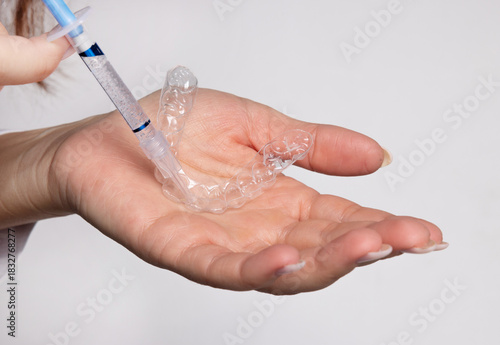 Woman holds whitening trays and solution on a minimal backdrop. Clear trays, dental model, case and syringes placed on the table. The woman applies gel into the trays for tooth care and bright smile.