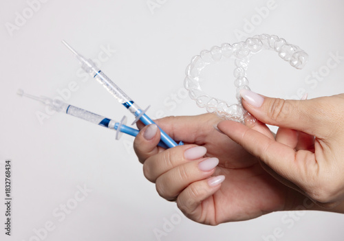 Woman holds whitening trays and solution on a minimal backdrop. Clear trays, dental model, case and syringes placed on the table. The woman applies gel into the trays for tooth care and bright smile.