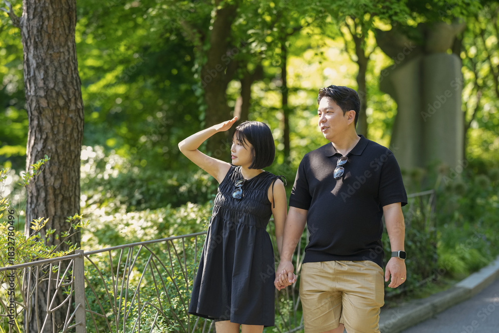 Fototapeta premium A man and woman are walking in a park, holding hands. The woman is wearing a black dress and the man is wearing a black shirt and khaki shorts. Scene is romantic and peaceful