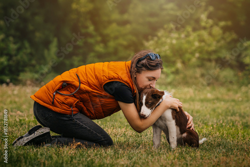 Woman Hugging Border Collie Puppy Outdoors. Young woman in an orange vest kneels on the grass and hugs her Border Collie puppy in a green park.