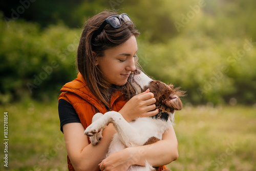 Woman Holding Border Collie Puppy in Park. Young woman in an orange vest gently hugs her Border Collie puppy while the dog nuzzles her face in a green park.