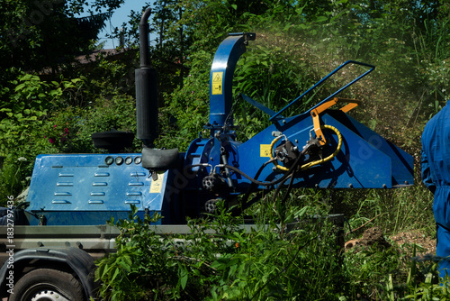 Male Arborist using a working wood chipper machine.The tree surgeon is wearing a safety helmet with a visor and ear protectors.
