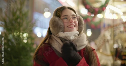 A young beautiful girl in a red coat walks in the evening at the European Christmas Market against the backdrop of a festive carousel with illuminations.