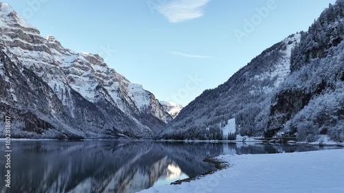 A serene winter look at Klöntalersee in Glarus, Switzerland, where a dark, motionless lake mirrors the steep, snow-draped Klöntal mountains beneath a crisp, clear blue sky.