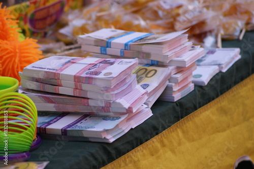 The photograph shows stacks of money neatly arranged on a table. The money is of various denominations, including 100- and 500-euro bills. Colorful jewelry and other objects are visible in the backgro