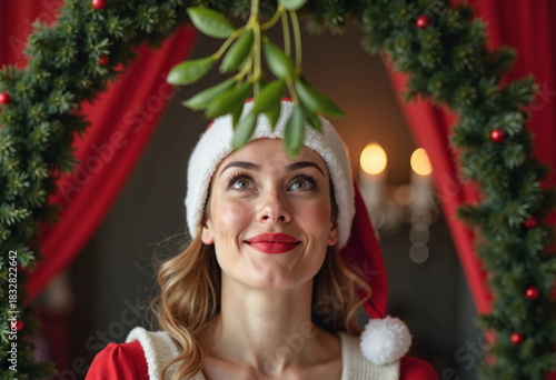 A woman wearing a red Santa hat and a red sweater smiles while holding green leaves above her head. 