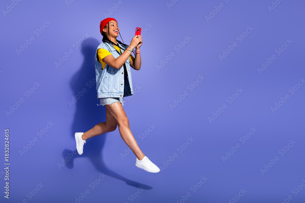 Obraz premium Joyful young woman leaps with phone in hand wearing denim streetwear against a violet background