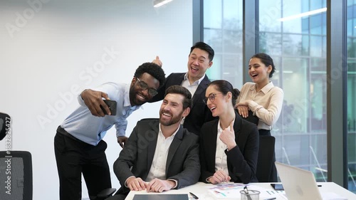 Group of happy diverse business people taking selfie using phone at workplace in business office. Smiling colleagues employees in formal wear taking joint photo sitting at desk in positive atmosphere
