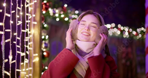 A young beautiful girl in a red coat walks in the evening at the European Christmas Market against the backdrop of illuminations and garlands.