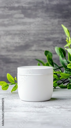 A white cosmetic jar is placed on a textured surface, with green leaves in the background. The studio shot has a neutral, clean aesthetic.
