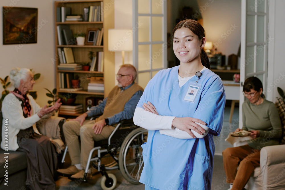 Obraz premium Young adult Hispanic woman nurse standing with arms crossed smiling at camera, senior Caucasian man in wheelchair and senior Caucasian women sitting and talking in background in care facility
