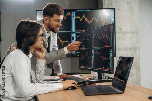 In formal clothes, by laptop and monitors. Two male exchange workers are in the office together