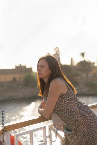 45-year-old woman on the railing of a ship gazing into the distance with a blurred background, behind her a village in Egypt 
