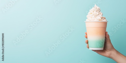 Hand holding colorful layered frozen drink topped with whipped cream against a pastel blue background, representing plant based refreshment and summer treats.