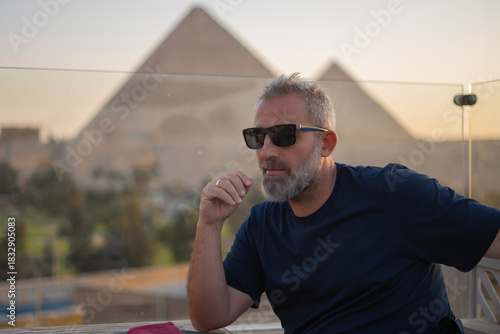 50-year-old man sitting on a terrace in front of the Pyramids of Giza in Cairo, Egypt, wearing a black T-shirt and sunglasses, an elegant and mature man.