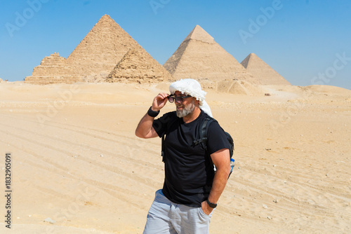 50-year-old man wearing a white turban or kufiya on his head, with a panoramic view of the entire Giza pyramids behind him. View of the Giza pyramids, middle-aged Caucasian man.