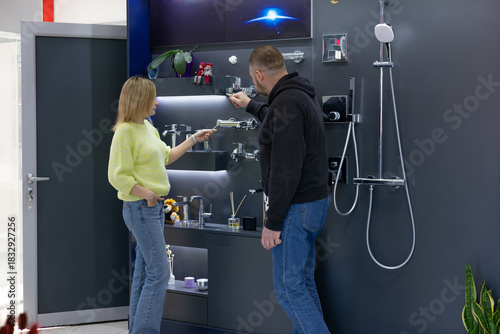 Plumbing store showroom. A consultant is explaining the features of various modern bathroom faucets and shower systems displayed on a dark wall to a male customer. Small business retail.