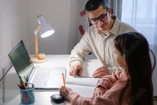 A father assists his young daughter with her homework at a brightly lit desk. They both appear engaged in the task, surrounded by school supplies and a laptop
