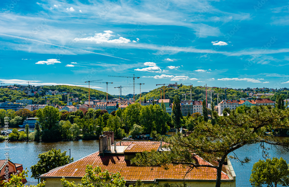 Obraz premium Construction cranes dominating Prague skyline over Vltava River.