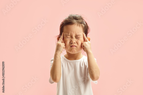 Quadro su tela Little Asian girl with tightly closed eyes presses her fingers to her temples, tries to remember something, expresses strong concentration, has forgotten something