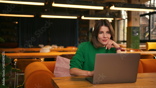 Young smiling freelancer woman working remotely on laptop in cafe or modern coworking