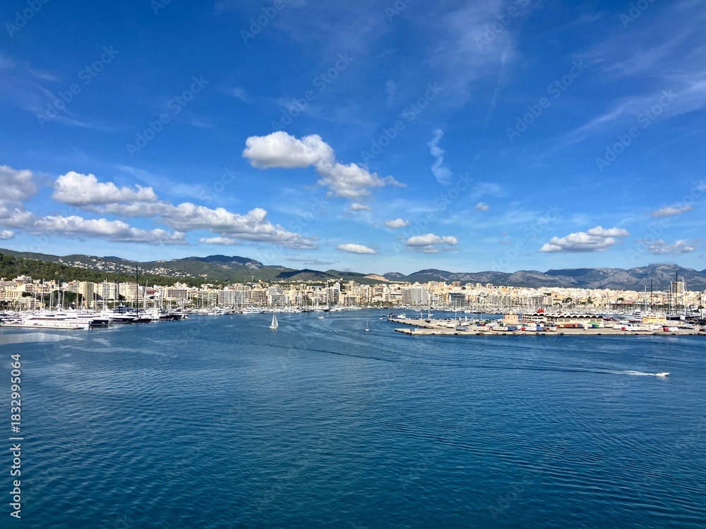 Fototapeta premium Scenic panoramic view of a coastal city with marina, yachts, and calm blue sea under a bright sky with scattered clouds, surrounded by hills and buildings