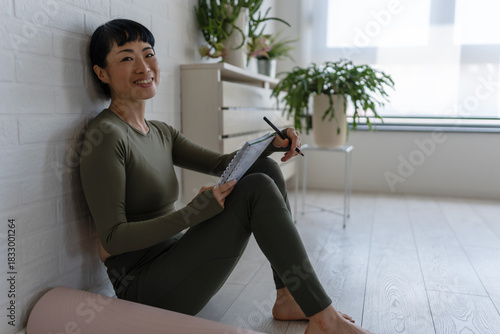 Smiling woman in sportswear sitting on floor at home with notebook, relaxing after workout and planning her fitness routine