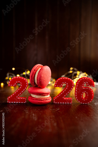 Red felt numbers 2026 and stacked red macarons with white filling celebrate the upcoming New year on a dark wooden background with bokeh lights, symbolizing sweetness and festivity
