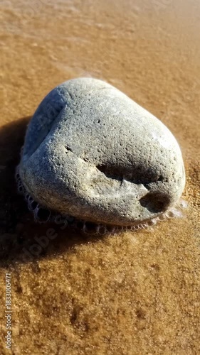A close-up view of a smooth, weathered stone resting on a sandy beach, where the edge of the water gently laps around its base.
