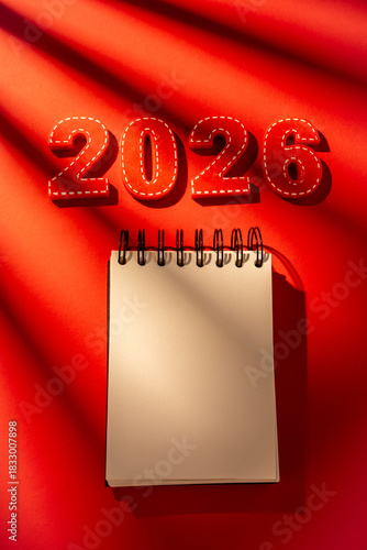 Red textured numbers 2026 featuring stitch details are positioned above a blank spiral notepad, symbolizing New year planning, resolutions, and future goal setting on a vibrant red background