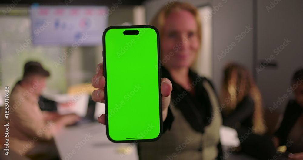 Obraz premium Vertical smartphone with green screen held by businesswoman in modern office, blurred coworkers collaborating in background with charts on digital screen