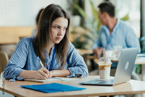 Female Student Taking Notes from Online Lecture in Sunny University Cafeteria During Lunch