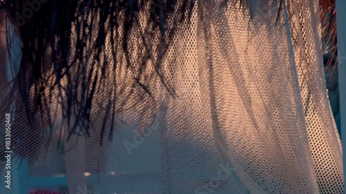 The hypnotic movement of a lightweight beach cabana canopy flutters in a tropical breeze under backlit sunlight. The fabric sways gracefully, creating a translucent silhouette with shimmering edges 